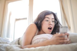 woman-lying-on-bed-holding-smartphone with shocked expression on her face